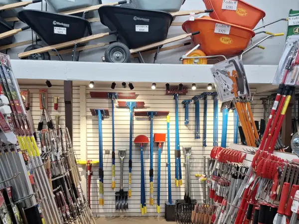 Tidbury's hardware store display features neatly arranged rakes, shovels, and tools hanging on a wall, with wheelbarrows stacked above. Bright lighting highlights the tools.