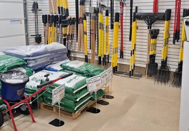 Tidbury hardware store section with neatly arranged tools. Shovels and rakes hang on a wall; bags of lawn products and a spreader are displayed. The scene is organized and practical.